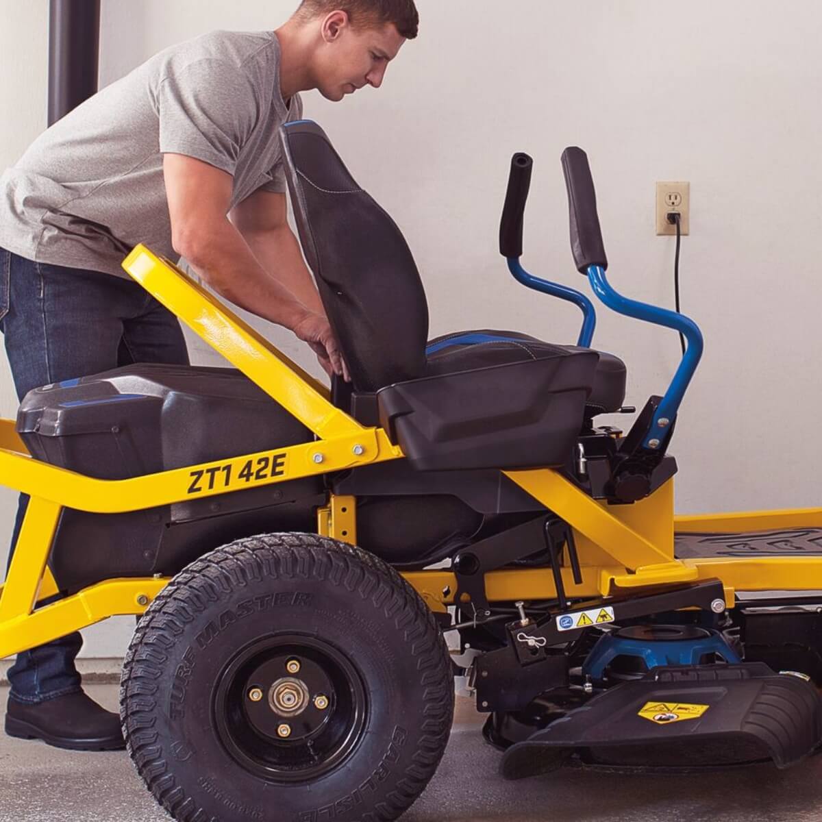 Shown from the side, a man adjusting a yellow and black ZT1 42E electric Cub Cadet ride lawn mower indoors.