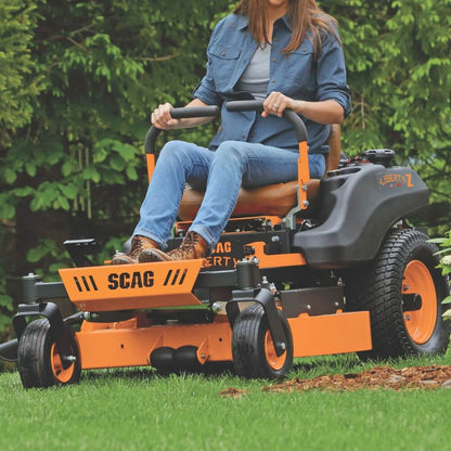 Person operating an orange SCAG Liberty 30 Inch riding lawn mower in a grassy area with trees in the background