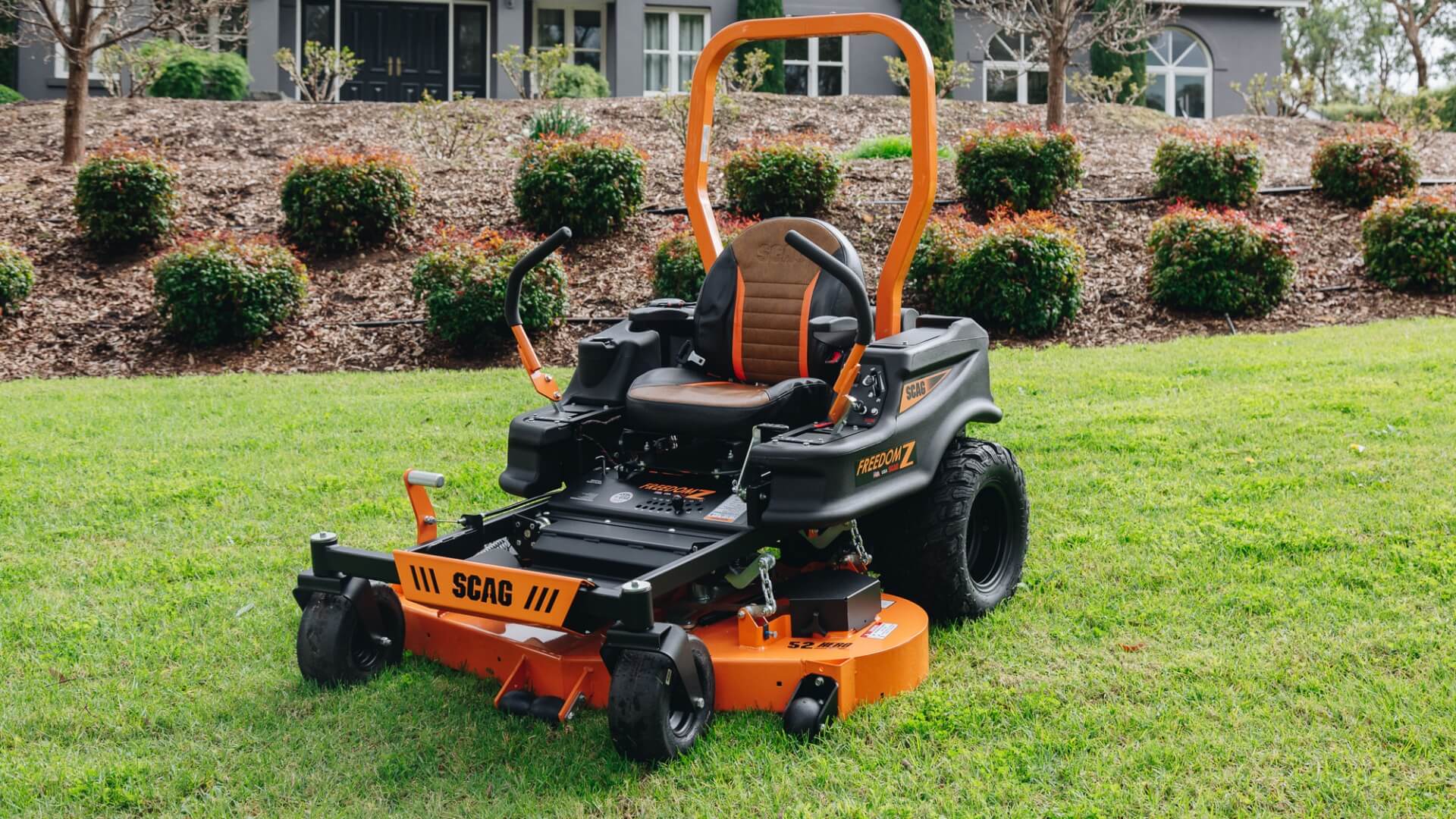 The SCAG Freedom Z ride on mower zero turn with roll over protection bar parked on green lawn with garden bed and large house in background.