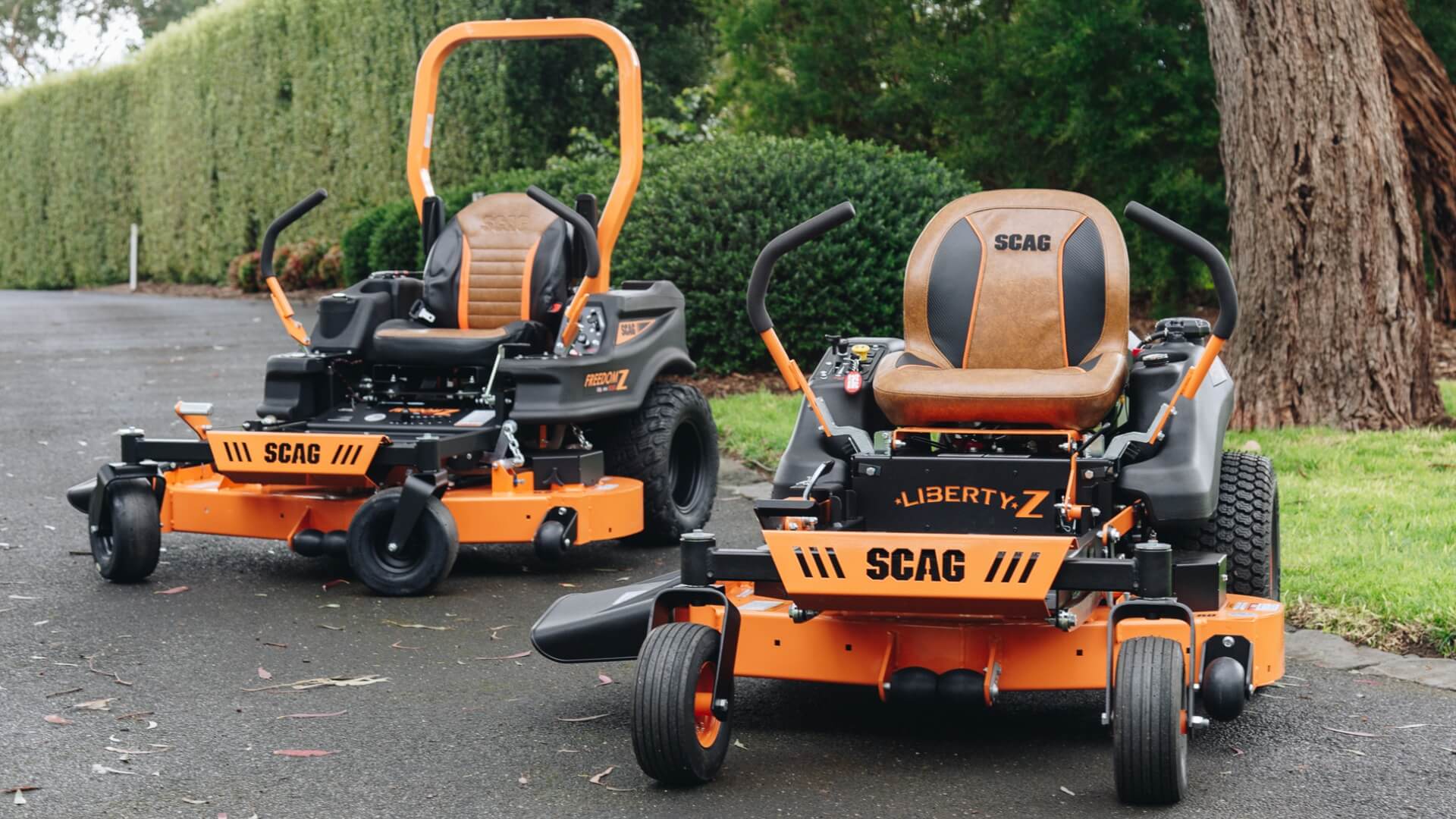 Two SCAG orange and black ride on mowers parked on a driveway with garden and trees.