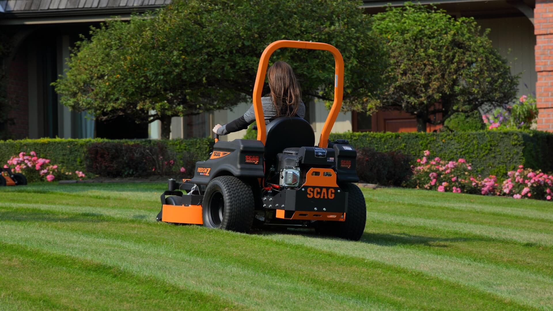 Womans back and long hair driving her orange and black Scag ride on mower across a beautiful green lawn with trees and house in the background.