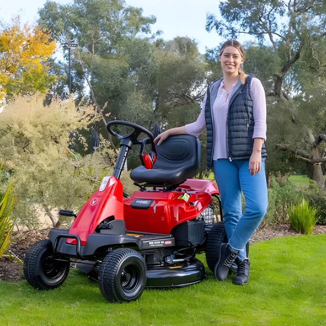 Woman wearing jeans and a vest smiling and looking at the viewer and standing next to her red Rover Mini ride on mower parked on a green lawn.