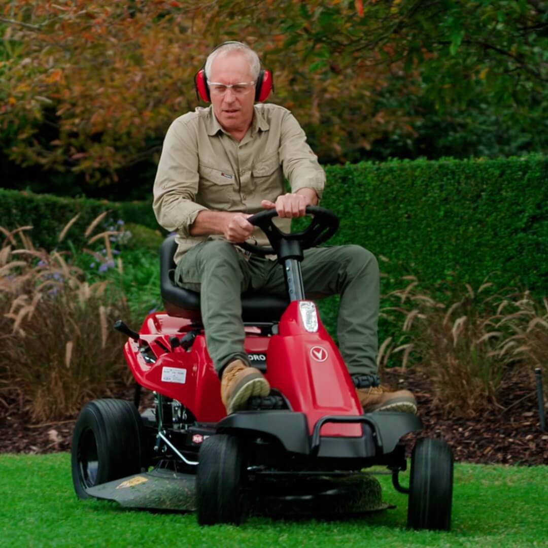 Man wearing gardening clothes and earmuffs steering his red Rover ride on mower to his right.