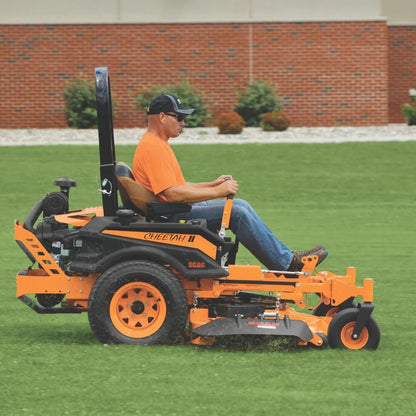 Man operating a large orange and black Cheetah SCAG lawn mower on grass with a brick wall in the background.
