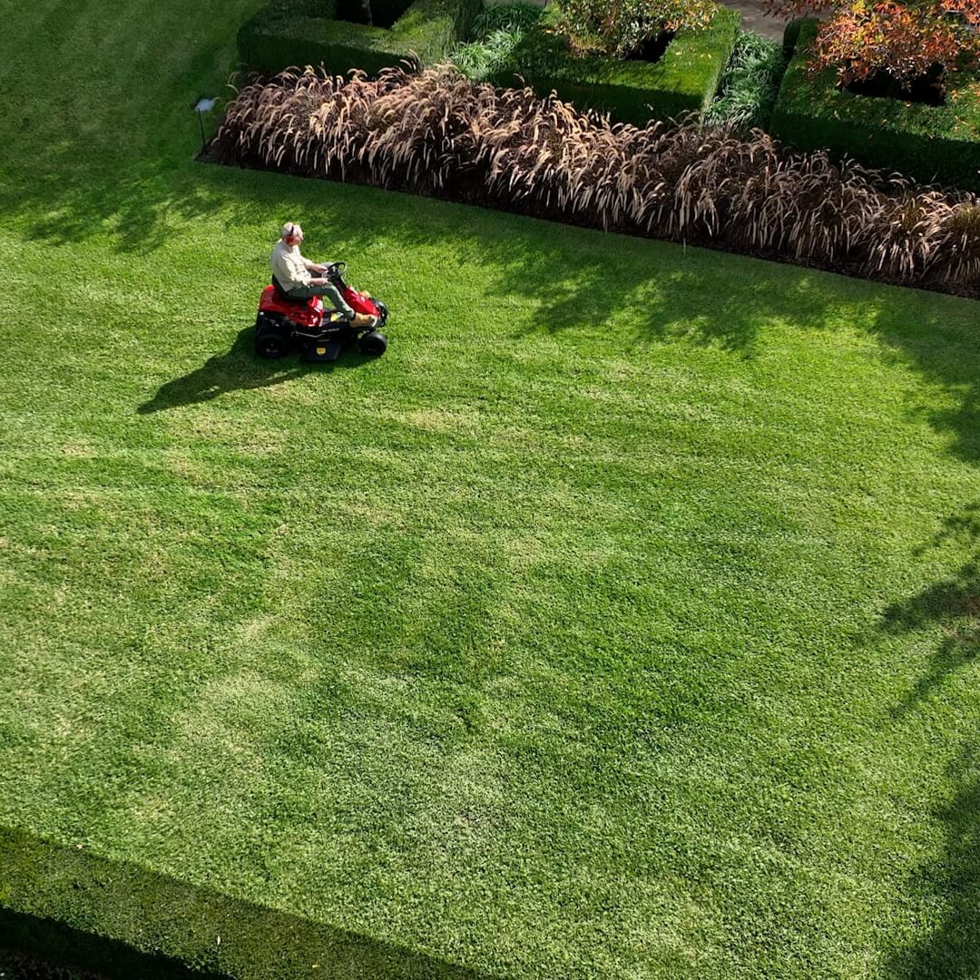 Person using a riding lawn mower on a green lawn with garden beds in the background.