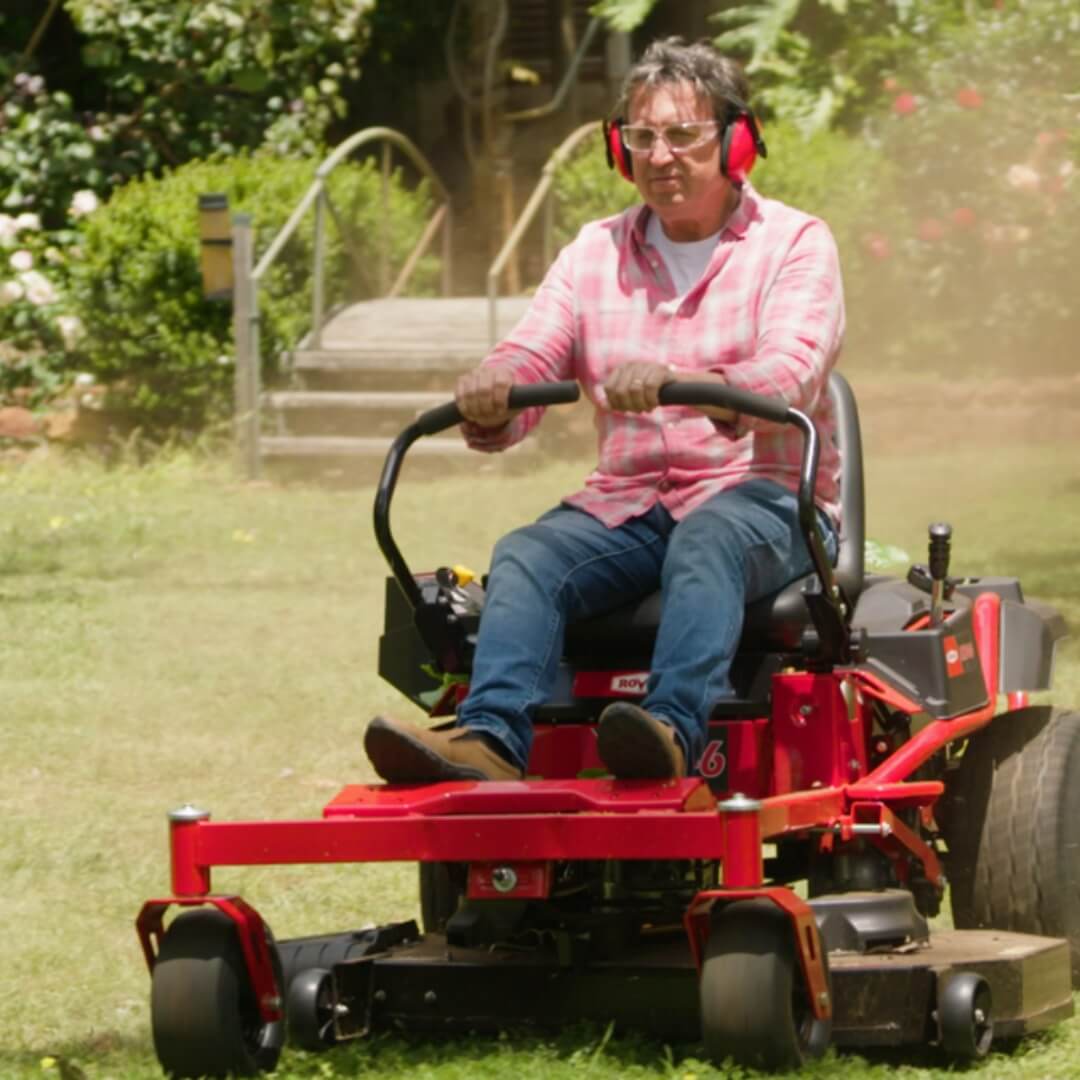 Man wearing earmuffs and steering his red Rover RZ ride on mower with lap bars.