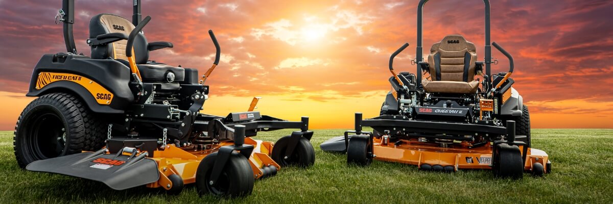 Two black and orange ride on mowers parked on green grass with bright orange sunset.