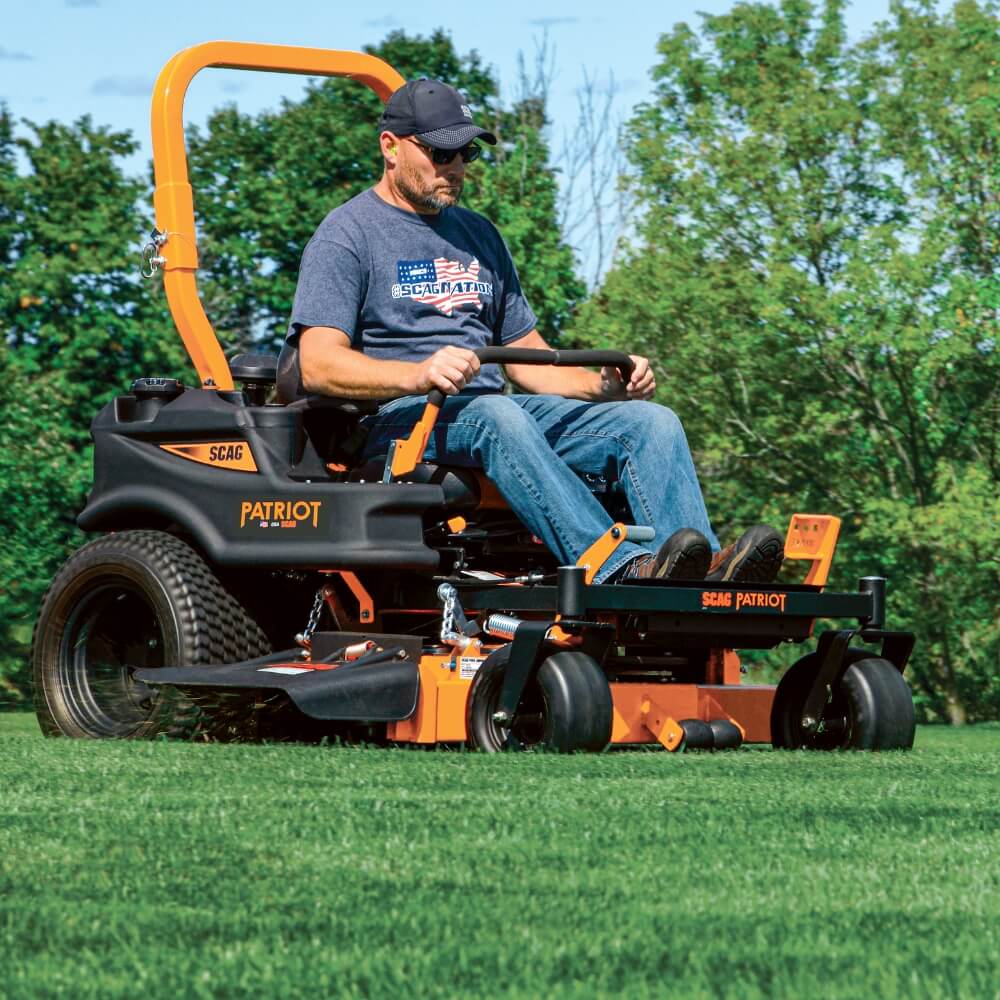 Man operating a SCAG Patriot riding lawn mower in a grassy area with trees in the background.