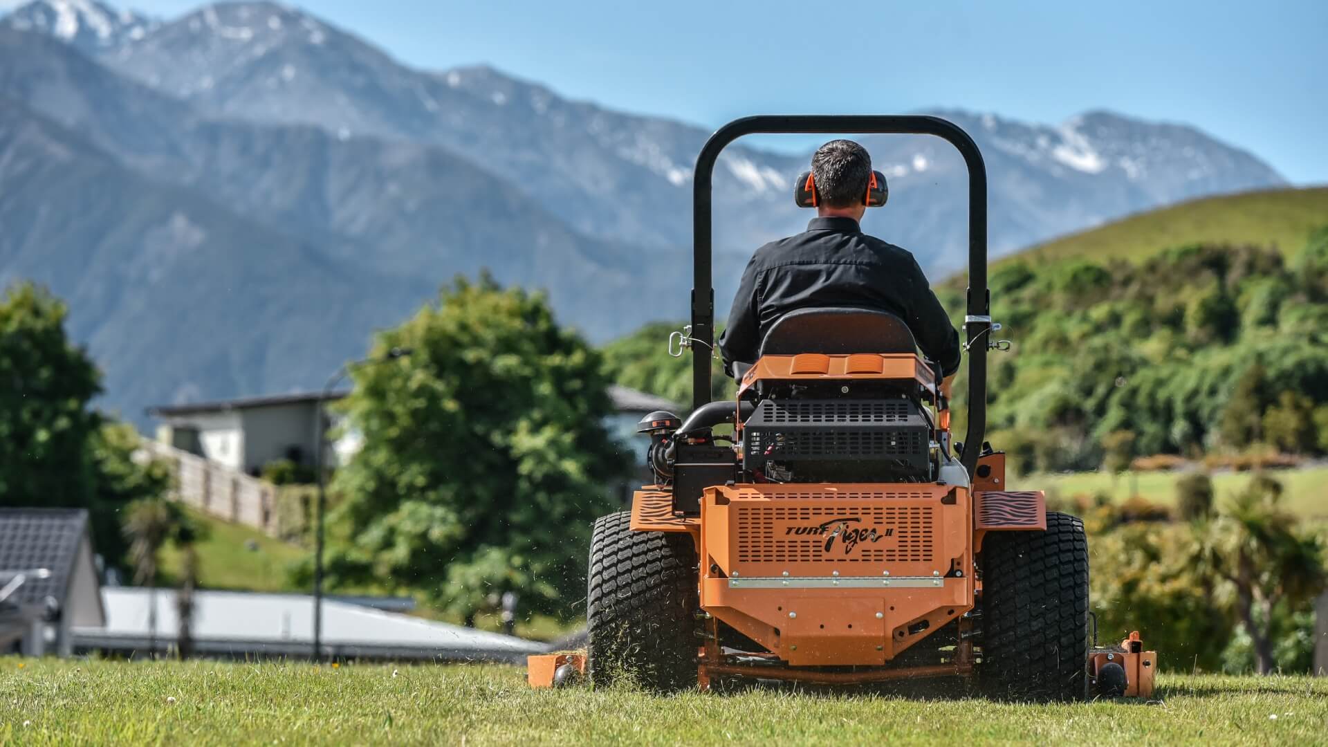 Man driving his Turf Tiger SCAG ride on mower across his property with green hills and snowcapped mountains in the far background.