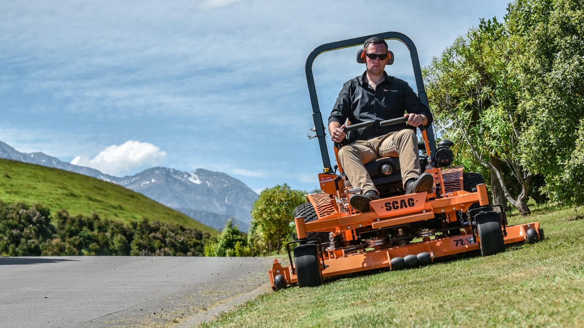 Man in black shirt wearing earmuffs mowing a sloping lawn alongside a road with green hills and snowy mountains in the far background.