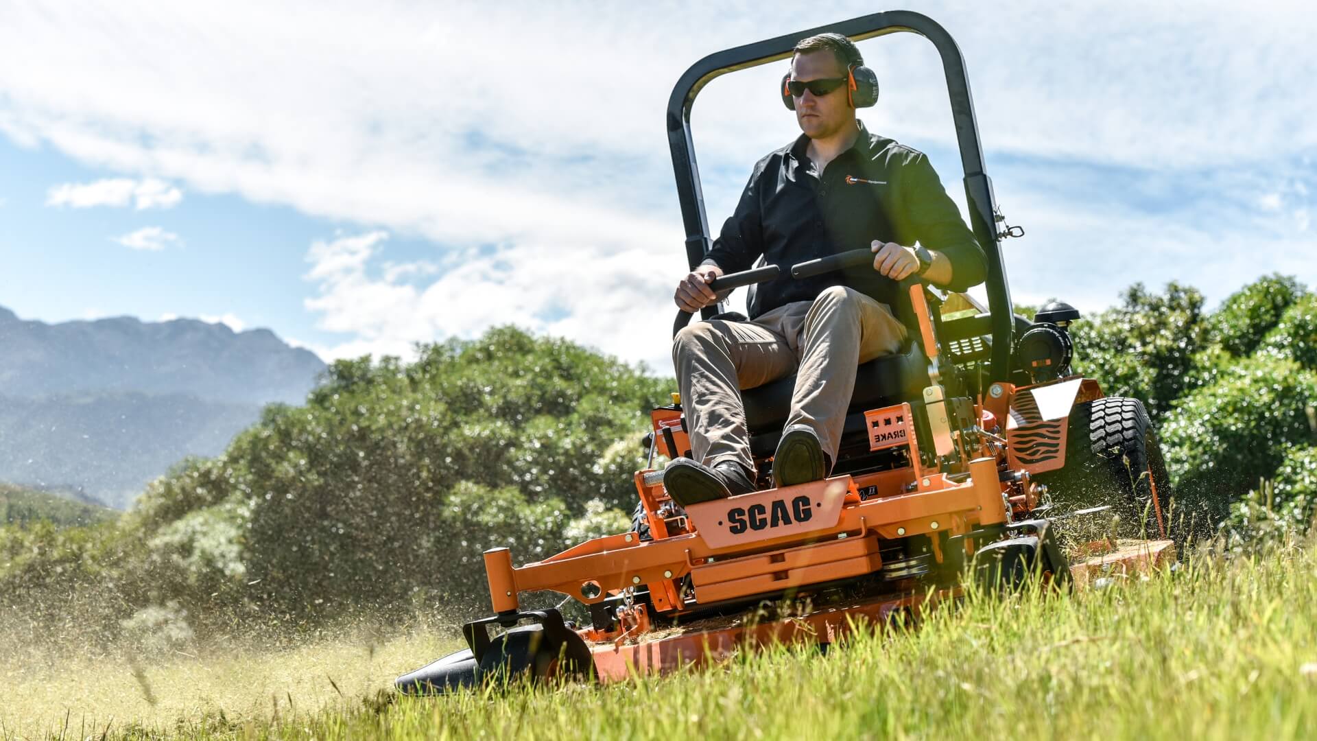 Main wearing black shirt and brown paints driving his SCAG ride on mower with lap bars cutting grass with trees and mountains behind him.