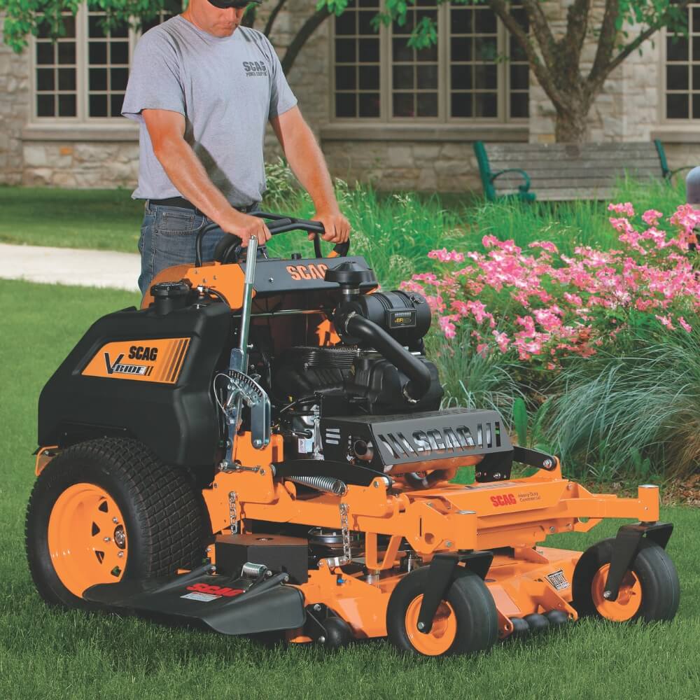 Person operating an orange SCAG mower in a garden with flowers and a building in the background