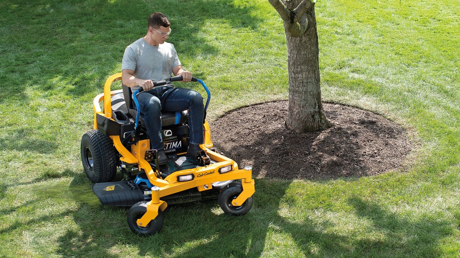 Man in grey t shirt driving his electronic yellow cub cadet ride on mower around a garden bed with tree.