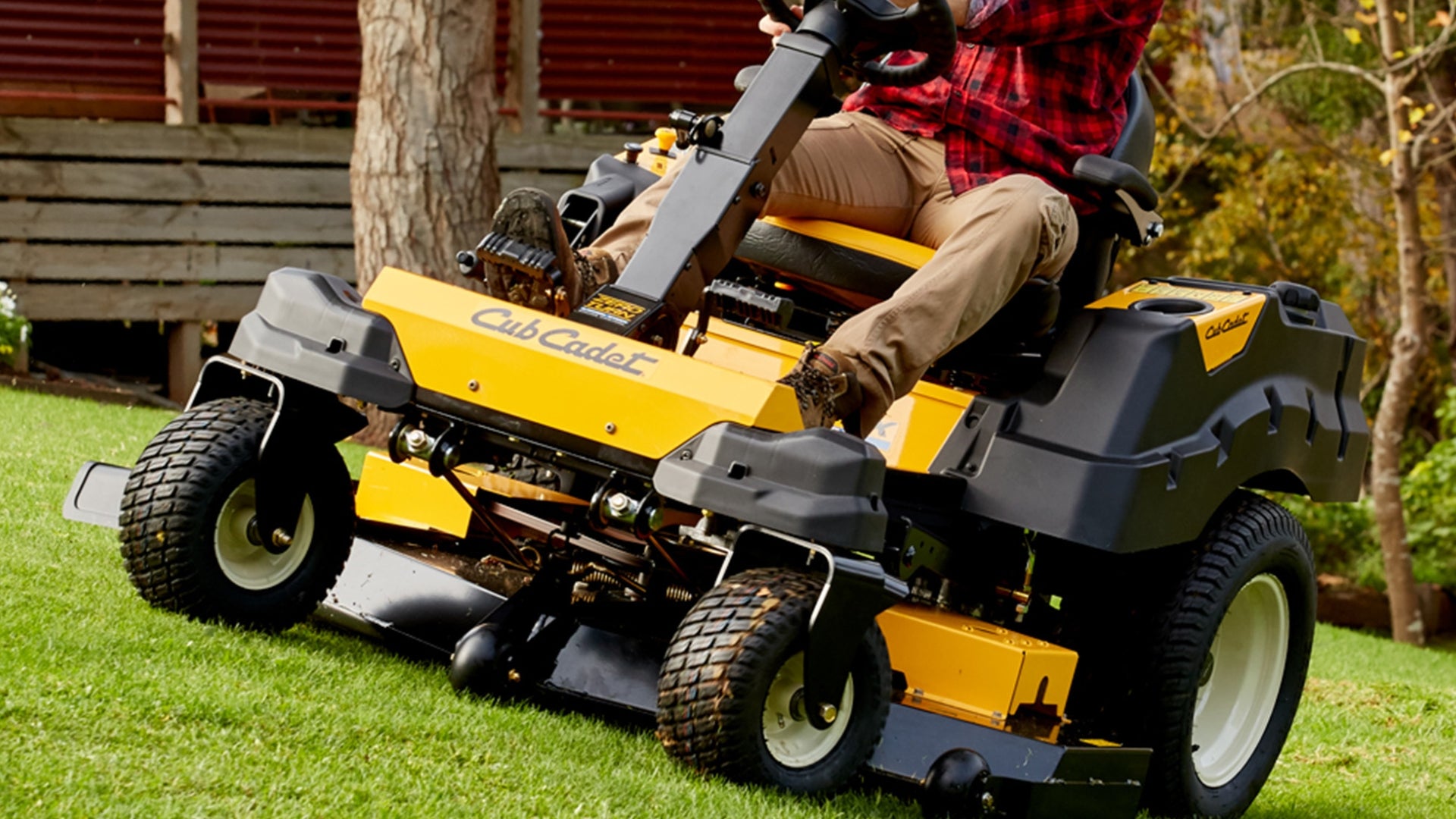 Driver of ride on mower navigating a sloping green lawn with house and trees behind.