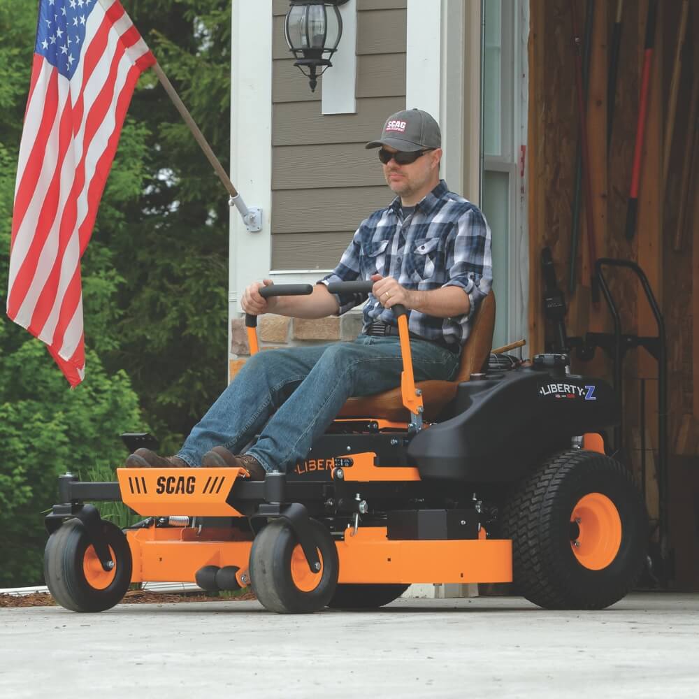 Man in cap driving his orange SCAG ride on mower out of his garage with house and American flag in background.