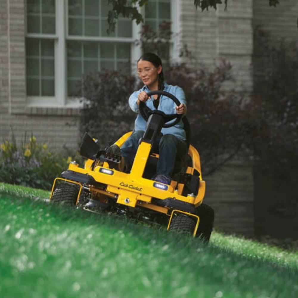 Woman steering her Cub Cadet ZTS1 ride on mower on a sloping grassed lawn with house in the background.