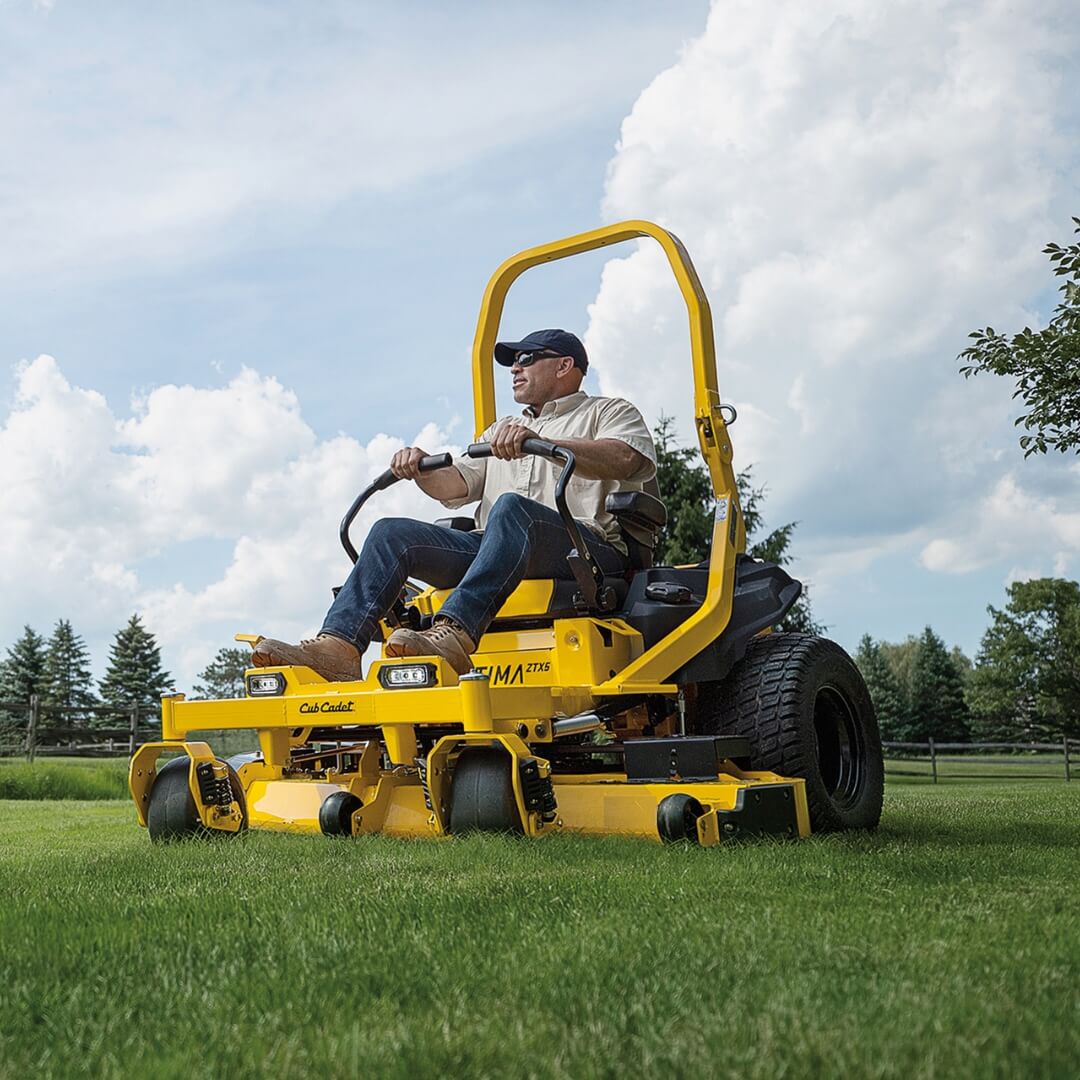 Man operating a yellow riding lawn mower on a grassy field with trees and blue sky in the background.